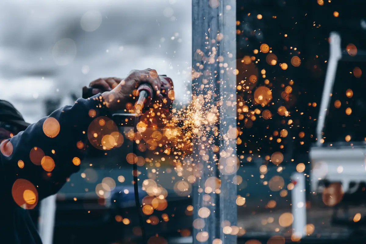 A close-up, shallow-depth-of-field shot of a worker's hands using a power grinder on a metal beam. Bright orange sparks fly toward the camera, creating a bokeh effect against a blurred industrial background.