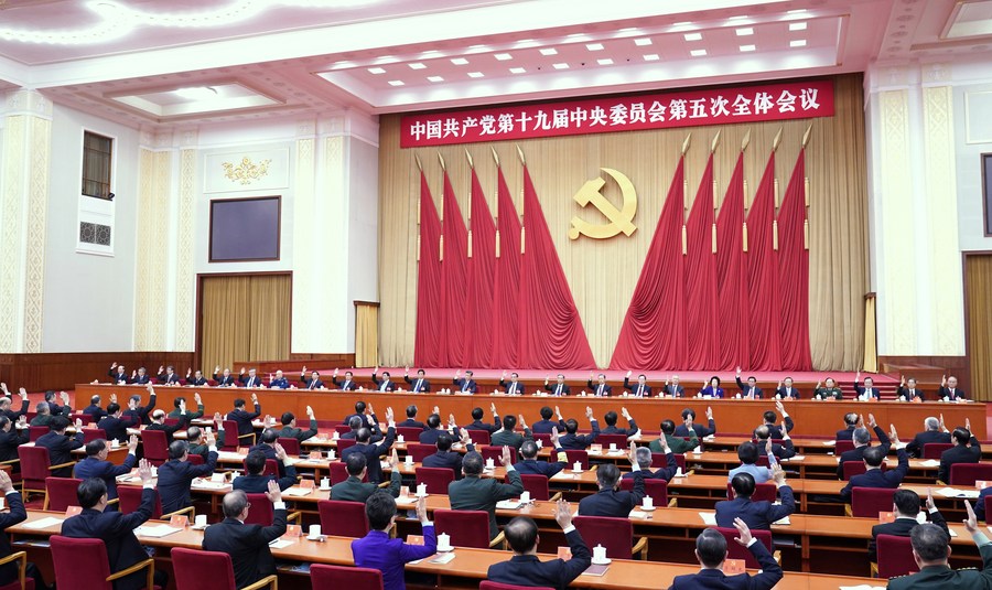 A wide-angle shot of a large assembly hall inside the Great Hall of the People in Beijing. Hundreds of delegates in formal attire are seated in rows of red chairs, raising their right hands in a unanimous vote. At the front, a long dais holds senior officials, backed by ten large red flags and a gold hammer and sickle emblem. A red banner at the top identifies the meeting in white Chinese characters.