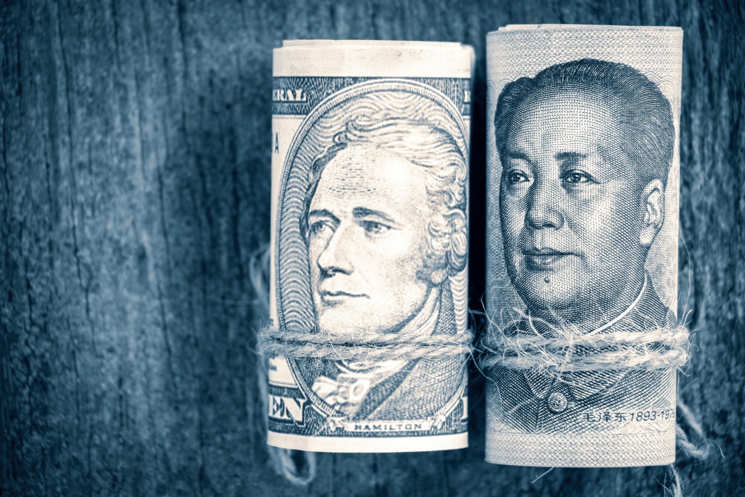 A close-up shot of two rolled-up banknotes standing side-by-side on a weathered wooden surface. On the left is a U.S. Ten-Dollar bill featuring Alexander Hamilton; on the left is a Chinese 100-Yuan bill featuring Mao Zedong. Both rolls are tied with rustic twine.