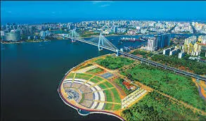 A wide aerial view of the Haikou Century Bridge in Hainan, China, showing its cable-stayed towers over the Haidian River, adjacent to a large coastal park and urban skyline.